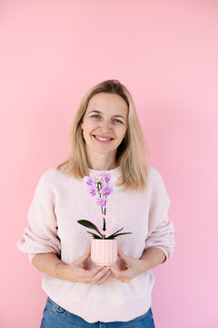 A blonde woman wearing a pink sweater is holding an orchid and standing in front of a pink background