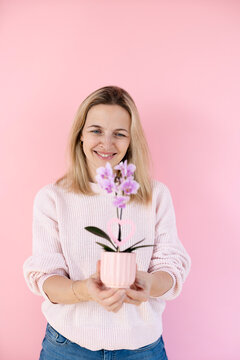 A blonde woman wearing a pink sweater is holding an orchid and standing in front of a pink background