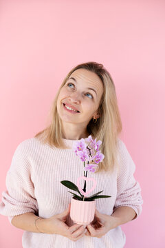 A blonde woman wearing a pink sweater is holding an orchid and standing in front of a pink background
