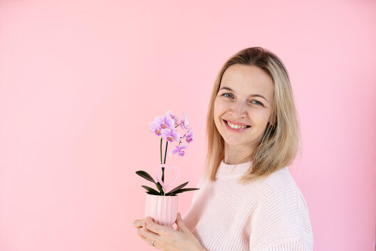 A blonde woman wearing a pink sweater is holding an orchid and standing in front of a pink background