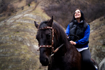 Fototapeta premium Happy woman and horse in nature. Low-angle view portrait of brunette female rider and black stallion outdoors in the mountains.
