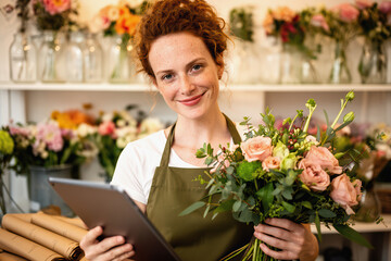 Fototapeta premium Female florist holding bouquet and tablet in a bright, busy flower shop.