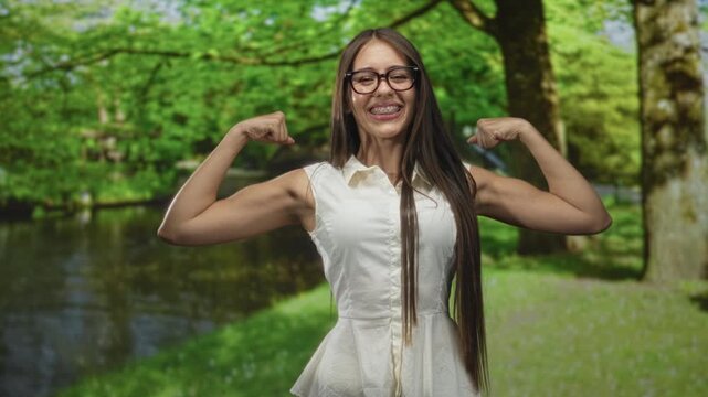 Teenage woman flexes bare biceps and smiles while posing in a forest by a pond, wearing white sleeveless blouse; confidence.