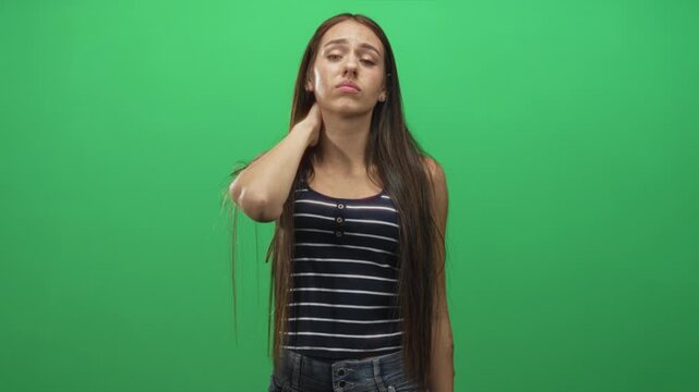 Young woman with long hair holds neck with hand, eyes closed and winces while standing in a green screen studio; discomfort recovery.