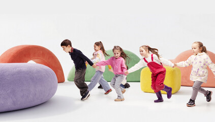 Naklejka premium Group of children holding hands running together on colorful abstract playground in studio. Concept of preschool activity, kids fashion advertising, active lifestyle promotion and childcare services.