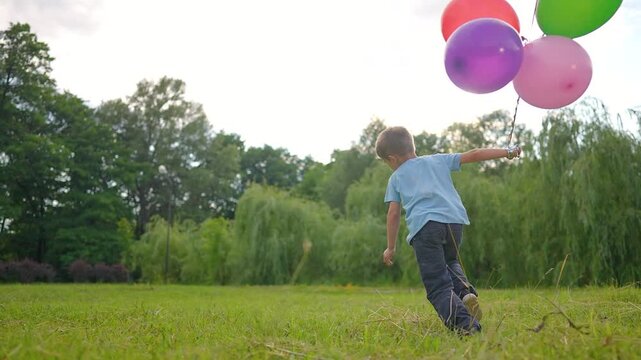 Happy boy runs with colorful balloons in park. Child playing outdoors with balloon bunch. Kid enjoying summer fun activity. Boy holds balloons while running. Child celebrates with balloons outside.