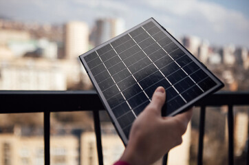 Fototapeta premium Hand holding a small solar panel against a balcony railing with a blurred cityscape in the background showcasing urban living and renewable energy solutions
