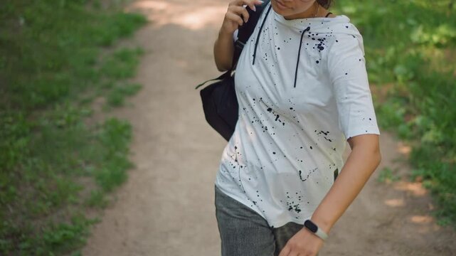 casual walker with backpack strap on leafy path, front torso shots show smartwatch on wrist, steady stride in white speckled hoodie, adjusts strap and reaches pocket while moving through dappled