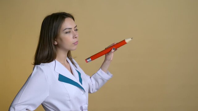 Young White woman adult girl in medical coat holding large pencil near face with thoughtful expression in studio. Concept of healthcare education, analysis and professional thinking