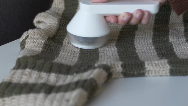 Hand gripping fabric shaver from above while removing lint from striped knit sweater at home. Clothing care technique suits sustainable fashion, conscious consumption. Second life for clothes