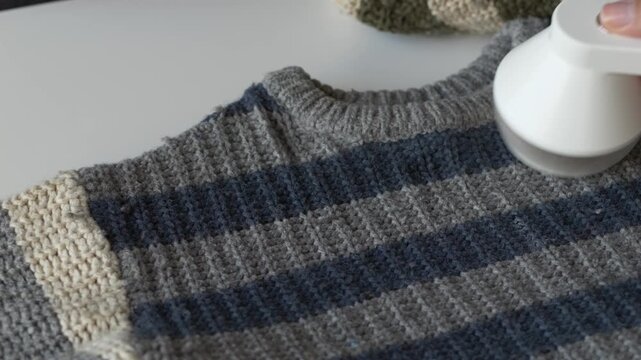 Hand using fabric shaver to remove lint from striped knit sweater on table. Clothing care process suits sustainable fashion, conscious consumption and garment maintenance lifestyle concept content.