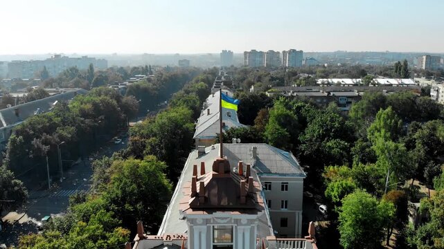 Aerial drone view of Sumy city center in Ukraine featuring historic architecture with a classical building topped by a Ukrainian flag, surrounded by green trees and urban streets.