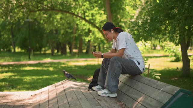 young man sitting on wooden roof scrolling smartphone, backpack beside him, dappled sunlight through trees, relaxed summer mood, candid park lifestyle scene showing concentration and quiet leisure