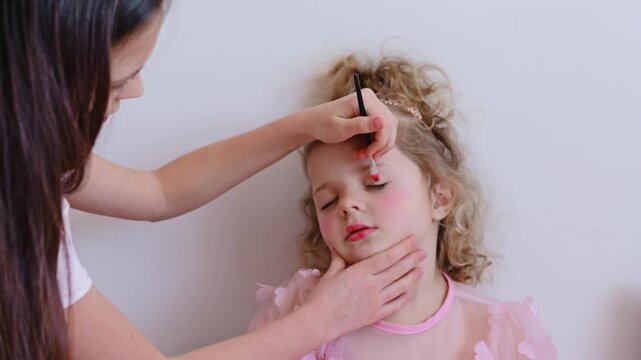 A young child sits while an older helper applies makeup on her face in a bright indoor space during daytime, showing a moment of fun and creativity