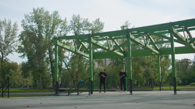 two athletes training under green pavilion, performing pullups on metal bars, urban park backdrop with trees and concrete, overcast sky, focused expressions, coach guiding partner, repetition
