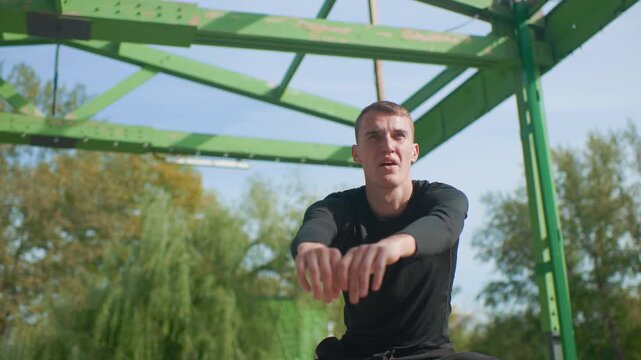 Outdoor trainer squats under green pavilion, focused athlete performing explosive squat and rowing motion, black longsleeve top, metal beams overhead, leafy park background, morning sunlight casting