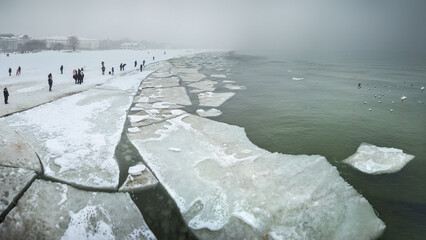 Winter landscape of a frozen Baltic Sea in Sopot, Poland, with ice floes on the water and people walking on the snowy beach under a foggy sky © ptk78