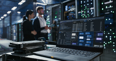 Laptop sits on a cart beside hard drives in data center.  Two people discuss network infrastructure...