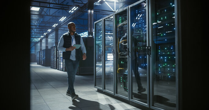 Data center IT specialist walks through aisles of server racks, using a tablet to inspect equipment, monitor network activity, and check connections in a cyber secure server room