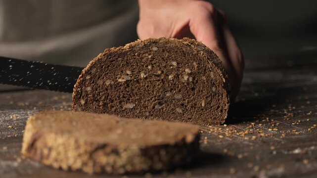 Baker slicing fresh loaf of rye bread with sesame seeds on a rustic wooden table in slow motion