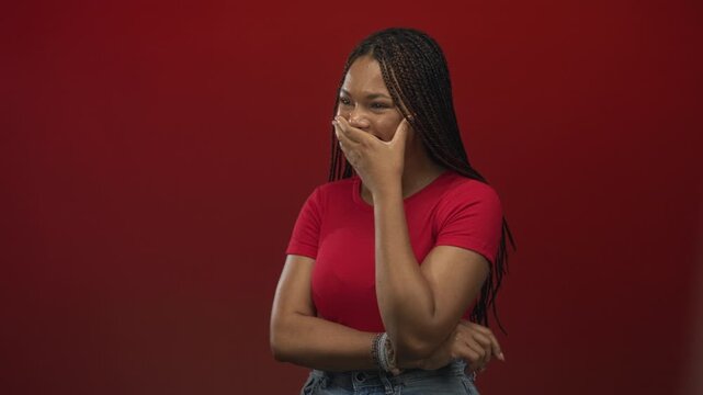 Woman covering mouth with hand, smiling and crossing arms in studio with deep red wall; lighthearted amusement.