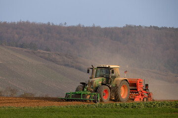 Fototapeta premium Tractor working on agricultural field with seeding machine in rural landscape at sunset