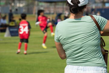 Fototapeta premium Asian parents watching and supporting their children playing football on a sunny afternoon at a local community tournament.