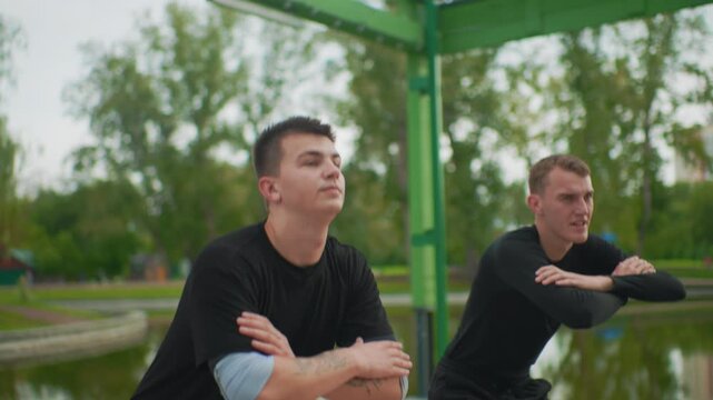 two white men standing by pond, park gazebo backdrop, stoic trainers warming up athletes, mentor advising recruit, coach observing stance, calm reflective water and green beams create disciplined mood