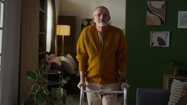Portrait of Caucasian senior man dressed in mustard-coloured buttoned cardigan and beige pants posing for camera standing with walking frame in living room