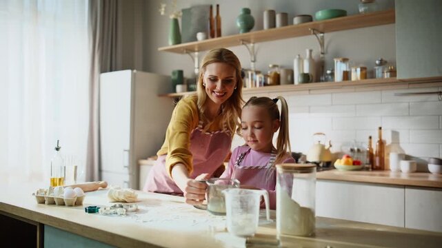 Tender mother helping daughter at kitchen. Child rolling dough mom sifting flour