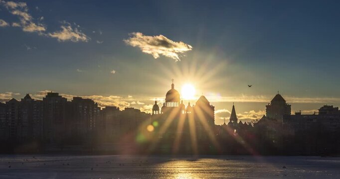 Bright sunset over Obolon embankment on the Dnipro River in Kyiv, Ukraine. Sun rays burst above church domes and skyline, reflecting on frozen water in a calm winter city scene.