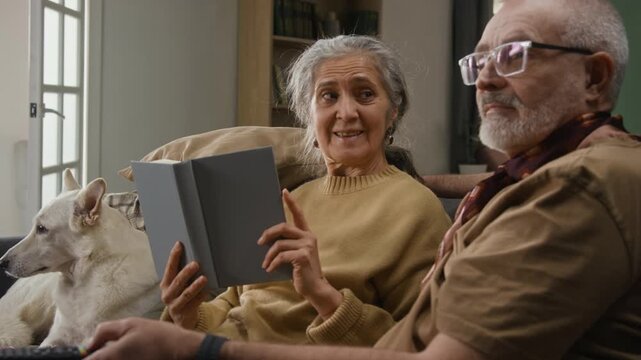 Senior man with glasses using TV remote, elderly woman reading book and looking at him with smile, white dog lying in background