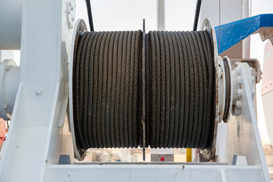 A heavy-duty steel cable winch on a ship's deck, showing thick wire rope neatly coiled around a large industrial drum, used for towing, mooring, or lifting operations in a maritime environment.