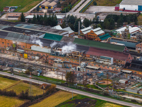 Aerial view of industrial buildings exhale plumes of white smoke against a backdrop of green fields, creating a stark contrast, Avezzano, Abruzzo, Italy.