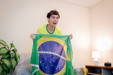 Torcedor brasileiro assistindo a um campeonato internacional de futebol pela TV em casa. Vestindo a camisa da seleção brasileira, ele torce com empolgação no sofá enquanto segura a bandeira do Brasil. © Marcio