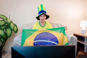 Torcedor brasileiro assistindo a um campeonato internacional de futebol pela TV em casa. Vestindo a camisa da seleção brasileira, ele torce com empolgação no sofá enquanto segura a bandeira do Brasil. © Marcio