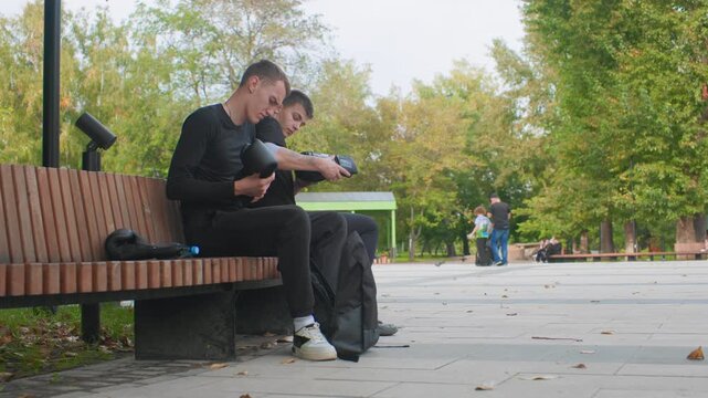 young caucasian man on bench, urban park autumn day, sitting alone with backpack, scrolling phone, head bowed, slumped posture, fallen leaves, distant pedestrians, soft overcast light, cinematic