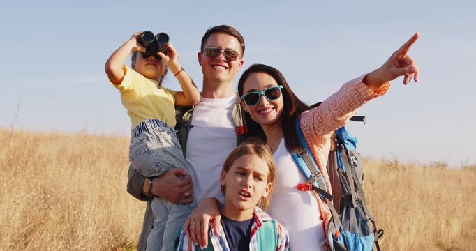 Family walking together in nature, happy outdoors. Parents and children pause on a meadow hike, pointing at sights as one uses binoculars. Celebrates joyful family outdoor adventure together.