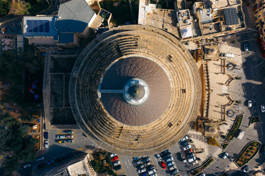 Aerial view of the Mosta Dome's majestic rotunda, a symphony of stone and shadow, crowned with an elegant cupola, amidst the bustling cityscape, Mosta, Malta.