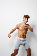 Fototapeta premium Muscular young man in light grey shorts with defined abs posing with arms raised on white background. Athletic body, youthful fitness, curly hair and confident expression, isolated studio portrait.