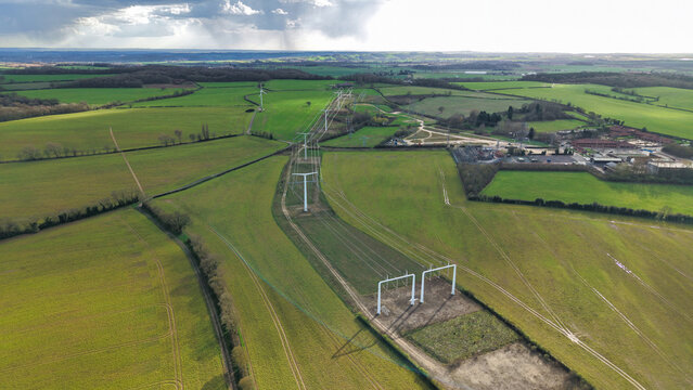 Aerial view of modern T-pylons marching across the verdant landscape, shadows dancing over the fields in stark contrast to the bright sky, Eakring, United Kingdom.