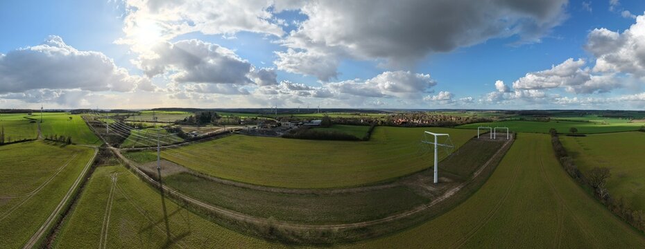 Aerial view of striking white T-pylons piercing the vibrant green fields under a vast sky of dramatic clouds, Eakring, United Kingdom.