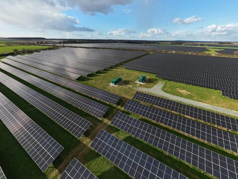 Aerial view of vast solar arrays glinting under a bright sky, juxtaposed against the verdant fields, presenting a stark contrast between technology and nature, Newark, United Kingdom.
