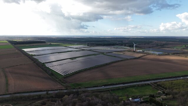 Aerial view of expansive solar panels glinting under a patchy sky alongside verdant fields and dark woods, Kirklington Road, Newark, United Kingdom.