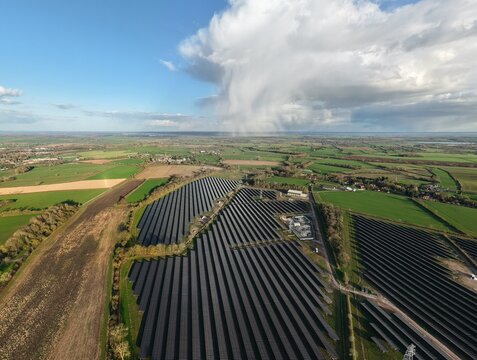 Aerial view of rows of solar panels glinting under the scattered sunlight, contrasting with the lush green fields and dramatic cloud formations, Halloughton, Southwell, United Kingdom.