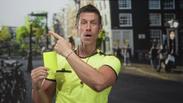 Man referee points finger while holding a yellow card on a city street outside buildings with bicycles and pedestrians, wearing neon shirt and whistle; surprise authority.