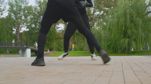 low angle park footwork, caucasian men moving. two athletes in black tracksuits practice rhythmic sparring steps on paved riverside path, sneakers scuffing brick, willow trees and bench in background,