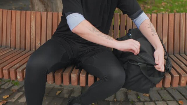 white man opening backpack on bench focused commuter hands unzip black pack revealing bottle and camera gear tattooed forearms wooden slatted bench cobblestone path daytime park scene