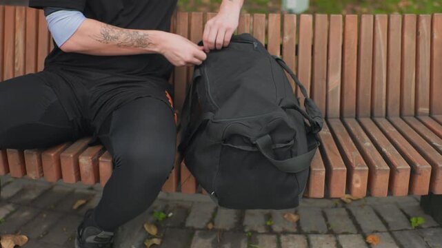 caucasian man retrieving inline skates from black duffel bag on wooden park bench, tattooed forearms, dressed in black activewear and leggings, cobblestone path and scattered leaves, hands pulling