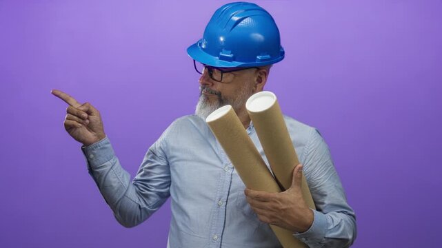 Man architect in blue hardhat holding rolled plans and pointing finger toward left in studio with purple backdrop; confidence planning.